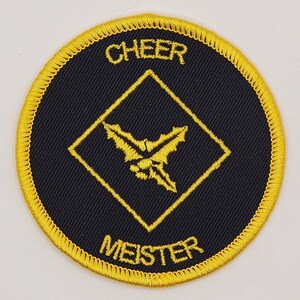 May include: A black and gold embroidered patch with a diamond shape in the center. The diamond has a yellow embroidered holly leaf design. The text "CHEER MEISTER" is embroidered in yellow around the diamond.