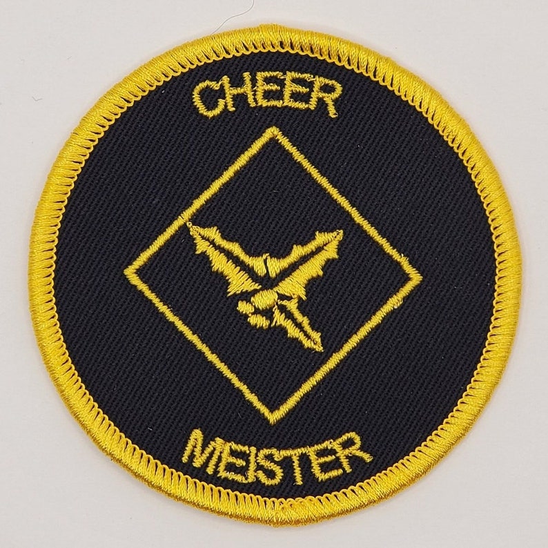 May include: A black and gold embroidered patch with a diamond shape in the center. The diamond has a yellow embroidered holly leaf design. The text "CHEER MEISTER" is embroidered in yellow around the diamond.