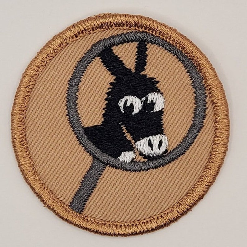 Op de afbeelding: Een bruine geborduurde patch met een grijze cirkel rond een zwarte cartoon-ezel met witte ogen en neus.