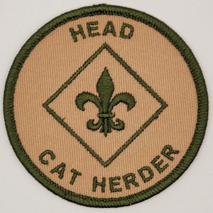 May include: A circular patch with a tan center and a green border. The patch features the words "HEAD" and "CAT HERDER" in green, surrounding a green fleur-de-lis emblem inside a diamond.