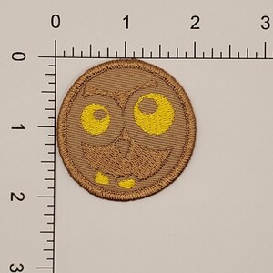 May include: A round, brown embroidered patch featuring a cartoon owl face with large yellow eyes and a smiling mouth. The patch has a stitched border and is likely designed to be ironed or sewn onto fabric. The design is simple and whimsical.