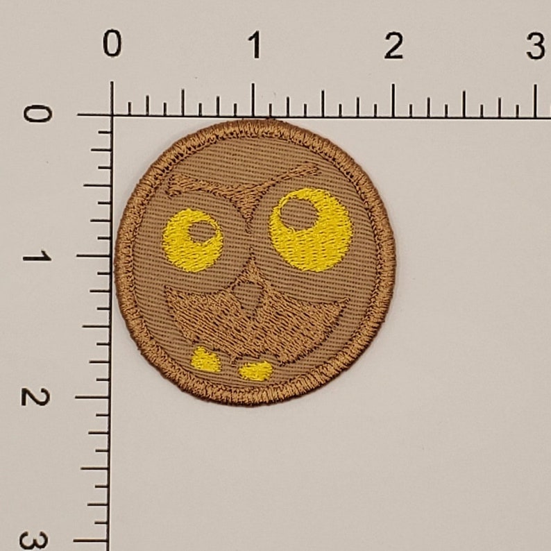 May include: A round, brown embroidered patch featuring a cartoon owl face with large yellow eyes and a smiling mouth. The patch has a stitched border and is likely designed to be ironed or sewn onto fabric. The design is simple and whimsical.