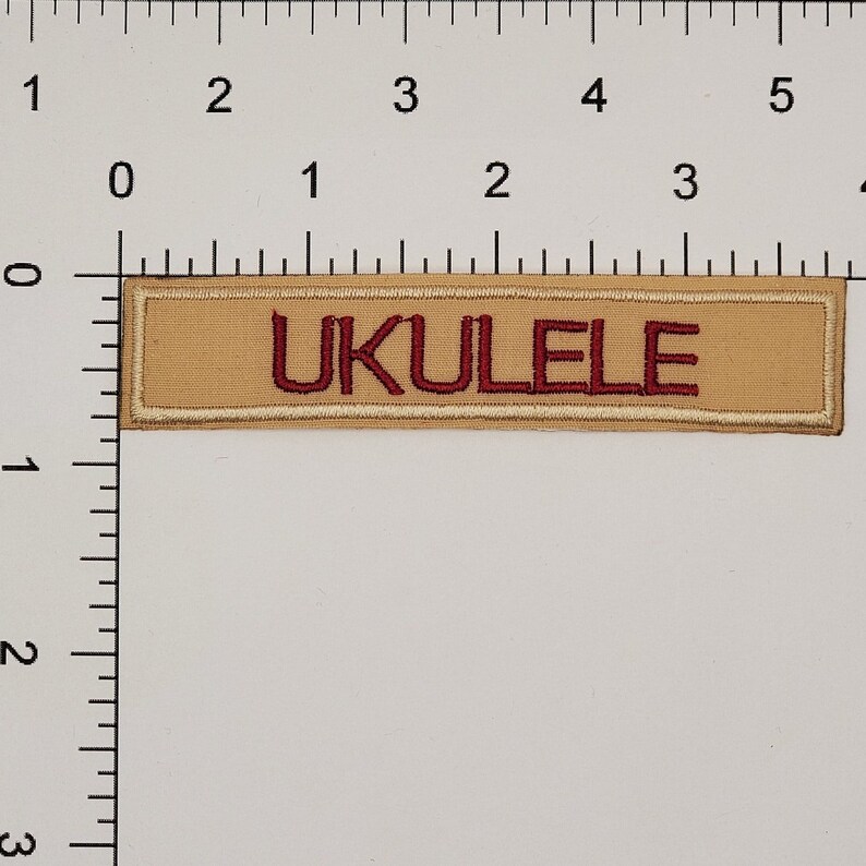 May include: Tan rectangular patch with the word "UKULELE" embroidered in burgundy thread. The patch has a tan border and is set against a white background. The patch is approximately 4 inches long.