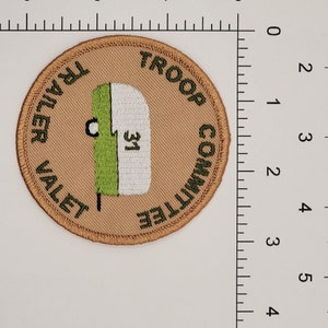 May include: A tan embroidered patch with green and white lettering. The text reads "Troop Committee Trailer Valet". A green and white illustration of a camper is in the center of the patch.