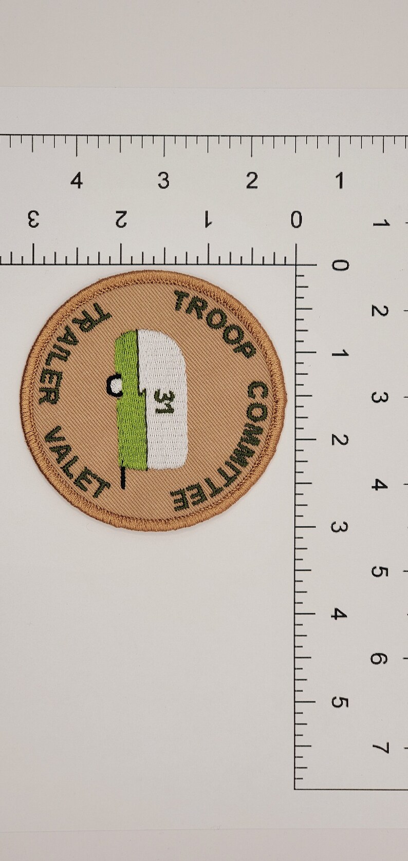 May include: A tan embroidered patch with green and white lettering. The text reads "Troop Committee Trailer Valet". A green and white illustration of a camper is in the center of the patch.
