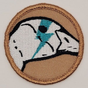 May include: A white embroidered patch with a black outline of a bird's head with a blue lightning bolt design. The patch is circular and has a brown border.