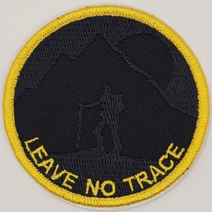 May include: Round black patch with a yellow border. The patch features a silhouette of a hiker with a walking stick in front of a mountain range. The words "LEAVE NO TRACE" are in yellow.