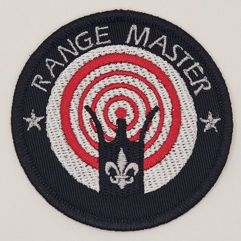 May include: Black embroidered patch with a white circle containing a red and white target with a black crown in the centre. The text "RANGE MASTER" is written in white around the circle.