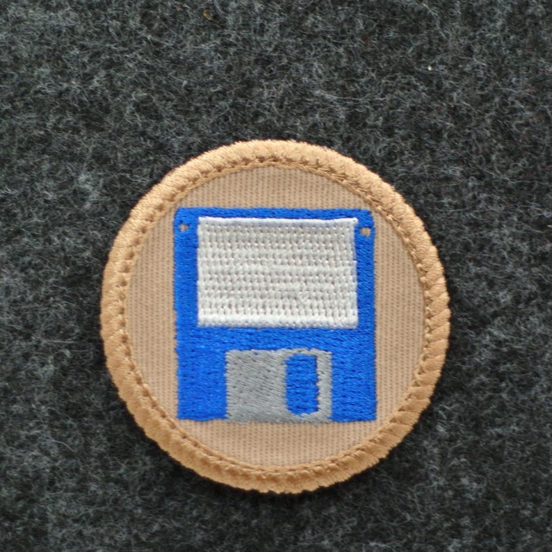 May include: A circular patch featuring a blue floppy disk design on a tan background. The floppy disk has a white textured area and a gray rectangular section. The patch is bordered with tan stitching and set against a dark gray fabric.
