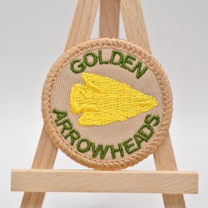 Pu&ograve; includere: Una toppa rotonda ricamata con le parole "GOLDEN ARROWHEADS" in verde sul bordo. La toppa presenta un disegno di punta di freccia gialla al centro, su uno sfondo beige, ed &egrave; incorniciata da un bordo beige.