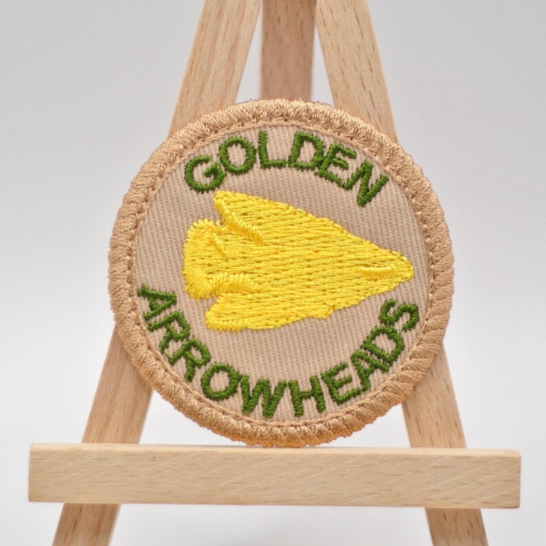 Pu&ograve; includere: Una toppa rotonda ricamata con le parole "GOLDEN ARROWHEADS" in verde sul bordo. La toppa presenta un disegno di punta di freccia gialla al centro, su uno sfondo beige, ed &egrave; incorniciata da un bordo beige.