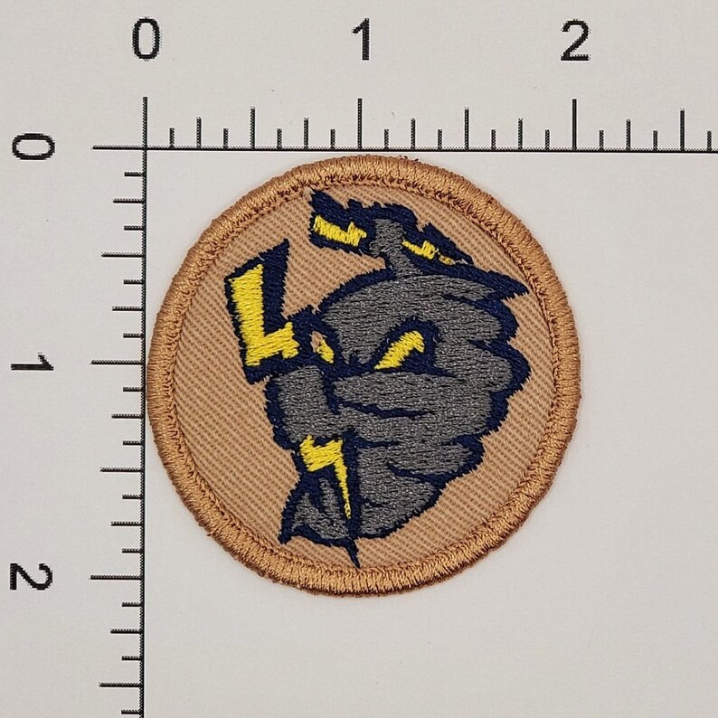 May include: Round embroidered patch featuring a gray and navy blue cartoon character with yellow lightning bolts. The patch is approximately 2 inches in diameter and has a tan border.