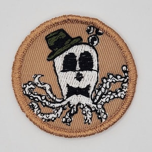 Puede incluir: Un parche bordado marr&oacute;n con un pulpo blanco que lleva un sombrero verde y una pajarita negra.