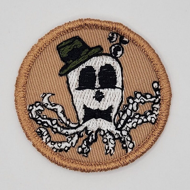 Puede incluir: Un parche bordado marr&oacute;n con un pulpo blanco que lleva un sombrero verde y una pajarita negra.
