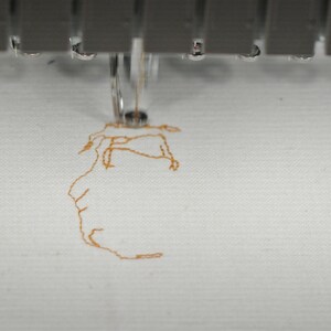 May include: Close-up of an embroidery machine in operation. The needle is stitching a detailed design in orange thread onto white fabric. The machine's metallic components are visible at the top, with water droplets.