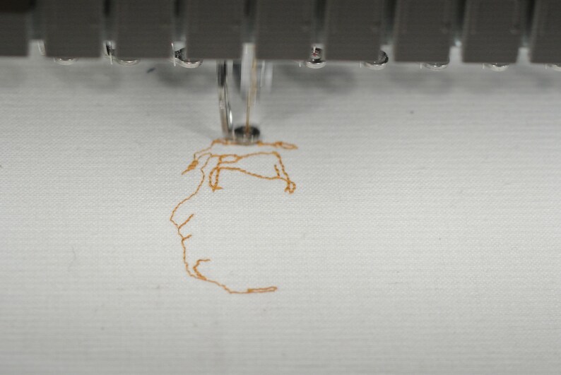 May include: Close-up of an embroidery machine in operation. The needle is stitching a detailed design in orange thread onto white fabric. The machine's metallic components are visible at the top, with water droplets.