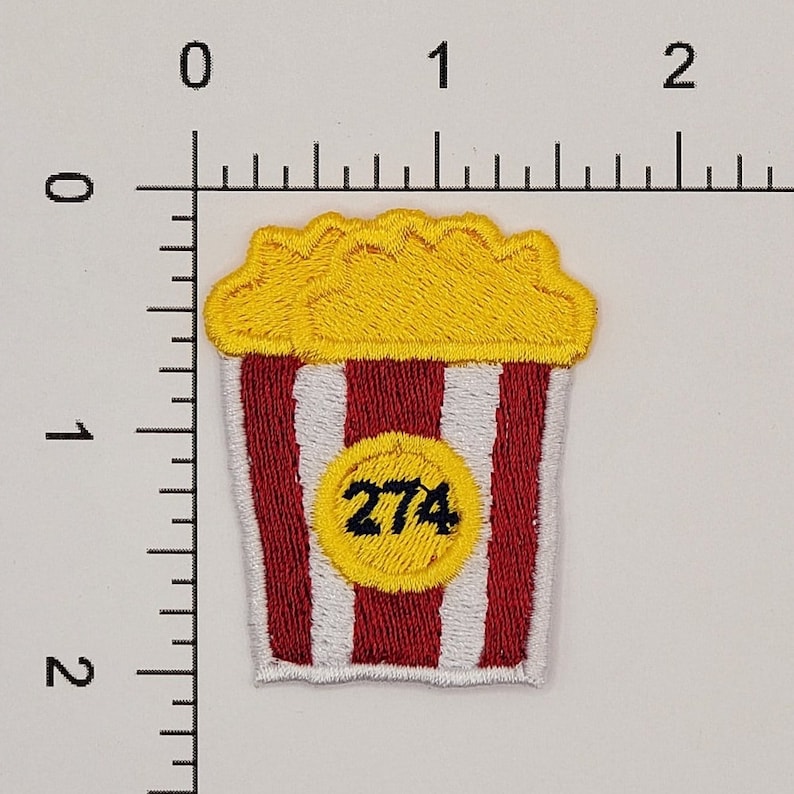 Peut inclure: Seau de popcorn &agrave; rayures rouges et blanches avec un cercle jaune contenant le num&eacute;ro 274. Le popcorn est jaune et a un bord festonn&eacute;.