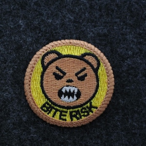 May include: Embroidered patch featuring a brown bear with an angry expression and visible teeth. The bear is set against a yellow background within a circular design, with the text "BITE RISK" in black. The patch has a brown border.