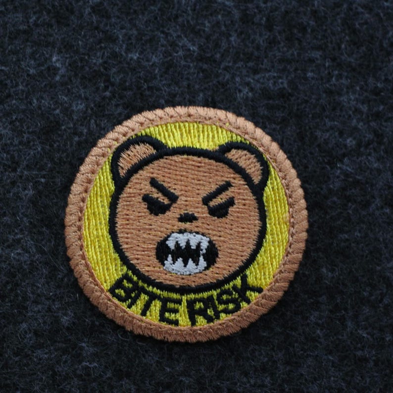 K&ouml;nnte beinhalten: Runder, gestickter Aufn&auml;her mit einem w&uuml;tenden braunen B&auml;ren, der seine Z&auml;hne zeigt. Der B&auml;r befindet sich auf einem gelben Hintergrund mit dem Text "BITE RISK" in Schwarz. Der Aufn&auml;her hat einen braunen Rand.