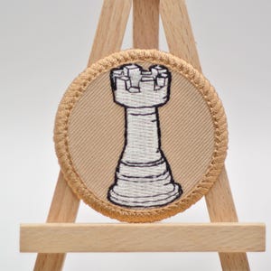 Op de afbeelding: Ronde geborduurde patch met een wit schaaktorenontwerp op een beige achtergrond. De patch heeft een beige rand en wordt weergegeven op een kleine houten ezel. De toren is een klassiek schaakstuk.