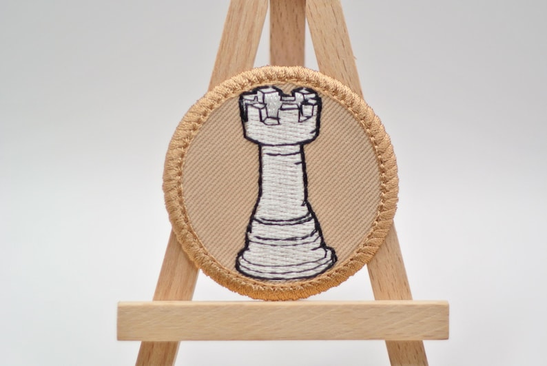 Op de afbeelding: Ronde geborduurde patch met een wit schaaktorenontwerp op een beige achtergrond. De patch heeft een beige rand en wordt weergegeven op een kleine houten ezel. De toren is een klassiek schaakstuk.