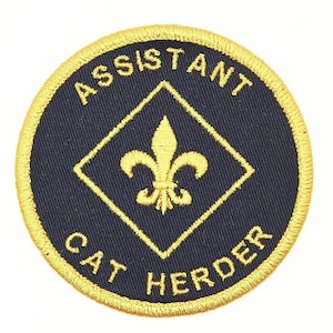 K&ouml;nnte beinhalten: Ein runder, gold gestickter Aufn&auml;her mit schwarzem Hintergrund. Der Aufn&auml;her tr&auml;gt die Aufschrift "Assistant Cat Herder" in einem Kreis um eine goldene Raute mit einer Fleur-de-lis in der Mitte.
