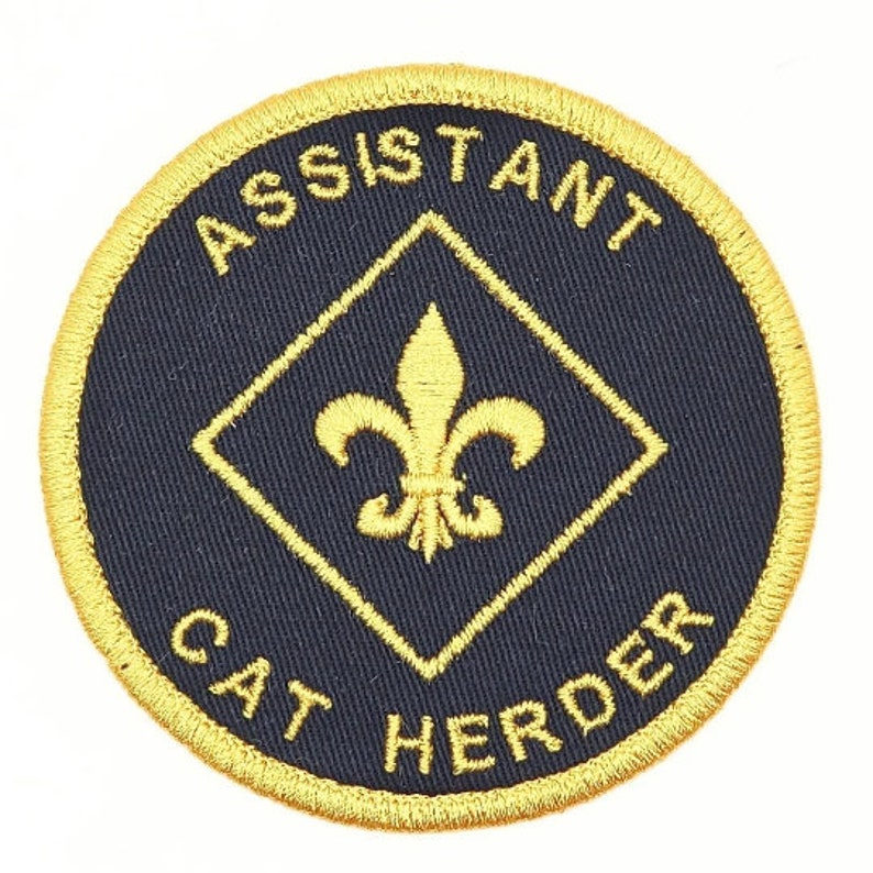 K&ouml;nnte beinhalten: Ein runder, gold gestickter Aufn&auml;her mit schwarzem Hintergrund. Der Aufn&auml;her tr&auml;gt die Aufschrift "Assistant Cat Herder" in einem Kreis um eine goldene Raute mit einer Fleur-de-lis in der Mitte.