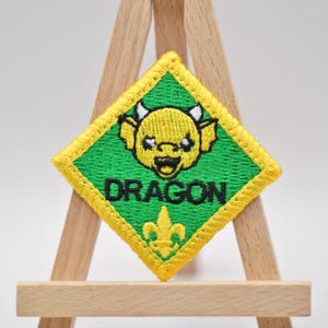 May include: Embroidered patch with a green background and a yellow border. The patch features a yellow dragon face with horns and the word "DRAGON" in black. A yellow fleur-de-lis is at the bottom.
