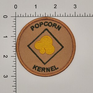May include: A round, tan fabric patch with a green diamond in the center. The diamond has a yellow popcorn kernel inside. The words "POPCORN KERNEL" are embroidered in green around the diamond.