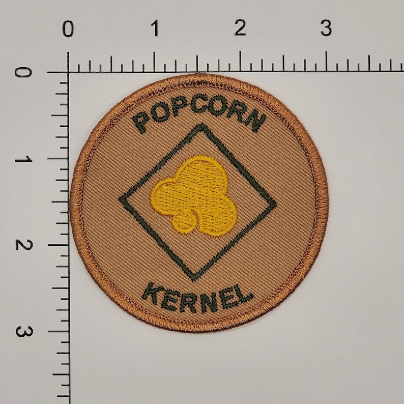 May include: A round, tan fabric patch with a green diamond in the center. The diamond has a yellow popcorn kernel inside. The words "POPCORN KERNEL" are embroidered in green around the diamond.
