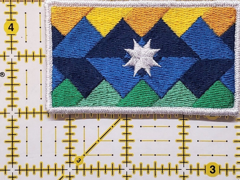 May include: A white embroidered patch featuring a blue, green and yellow mountain design. A white star is in the centre of the patch.