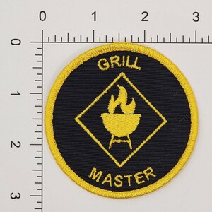 Peut inclure: Un &eacute;cusson brod&eacute; jaune et noir avec le texte "GRILL MASTER" et un losange jaune avec un grill et des flammes &agrave; l'int&eacute;rieur.