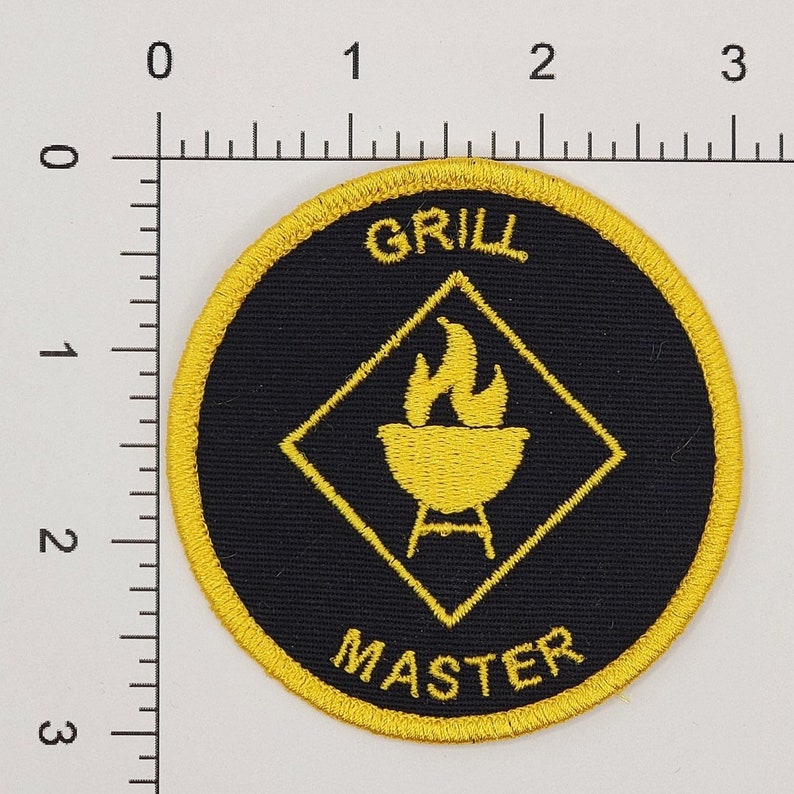 Peut inclure: Un &eacute;cusson brod&eacute; jaune et noir avec le texte "GRILL MASTER" et un losange jaune avec un grill et des flammes &agrave; l'int&eacute;rieur.