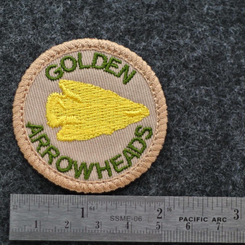 Pu&ograve; includere: Una toppa circolare con le parole "GOLDEN ARROWHEADS" ricamate in verde su sfondo beige. Una freccia gialla &egrave; al centro. La toppa &egrave; bordata con filo beige e si trova su una superficie grigia.