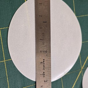 May include: A light gray, circular object is measured with a metal ruler. The ruler shows the diameter of the circle is approximately 4 inches. The object is on a green cutting mat.
