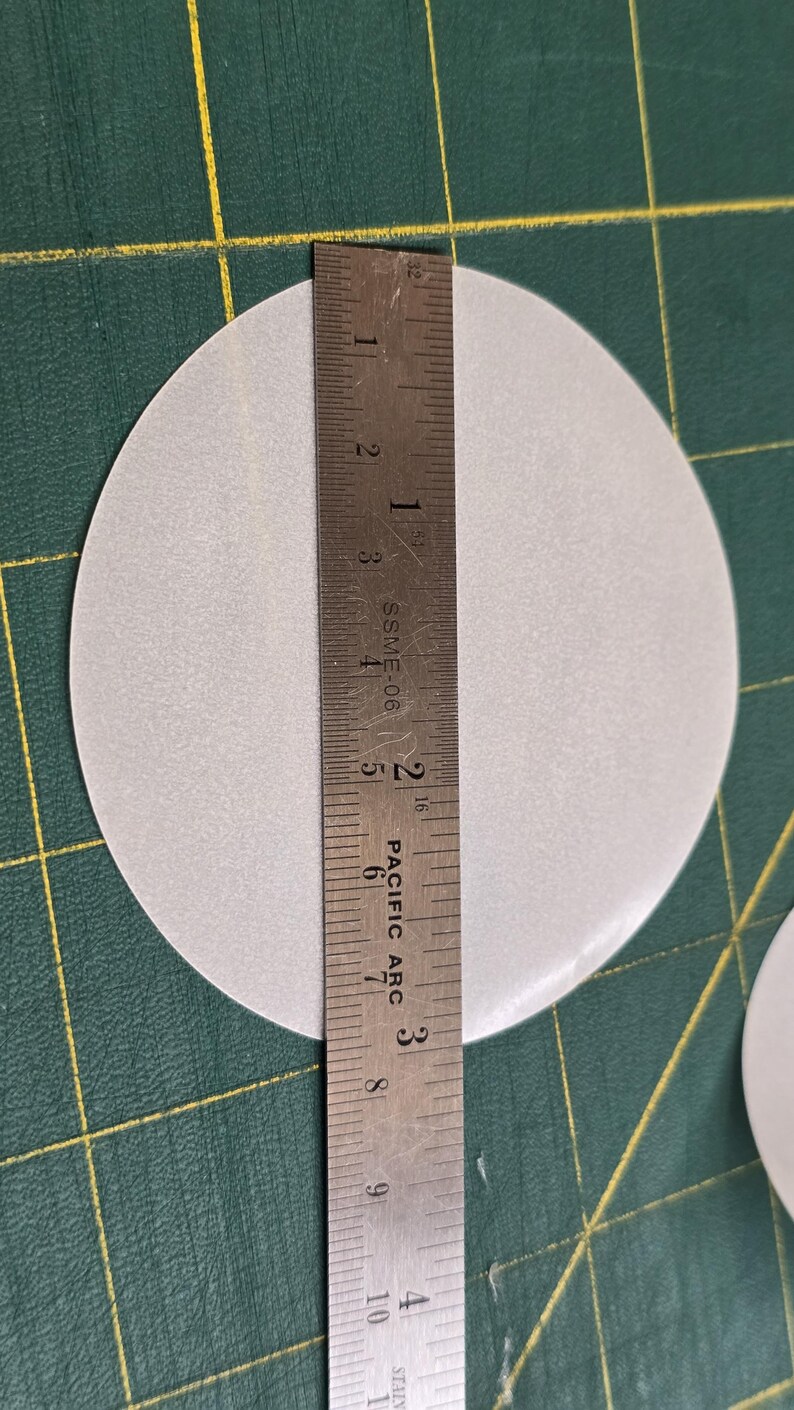 May include: A light gray, circular object is measured with a metal ruler. The ruler shows the diameter of the circle is approximately 4 inches. The object is on a green cutting mat.