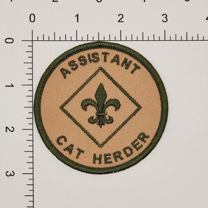 May include: A tan and green embroidered patch with the text "Assistant Cat Herder" and a green fleur-de-lis in a diamond shape.