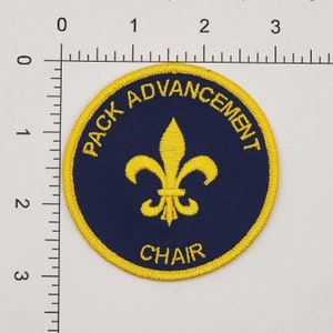 May include: A round, yellow embroidered patch with a blue background. The patch says "Pack Advancement Chair" with a fleur-de-lis in the centre.