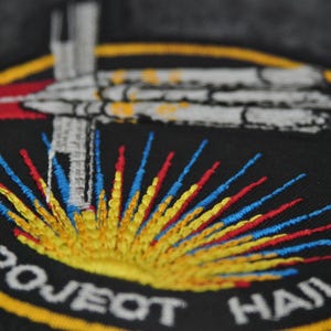 Puede incluir: Parche bordado con fondo negro, con un dise&ntilde;o de nave espacial y el texto "PROJECT HAIL MARS". El parche tiene un borde amarillo y detalles coloridos en rojo, azul y amarillo.