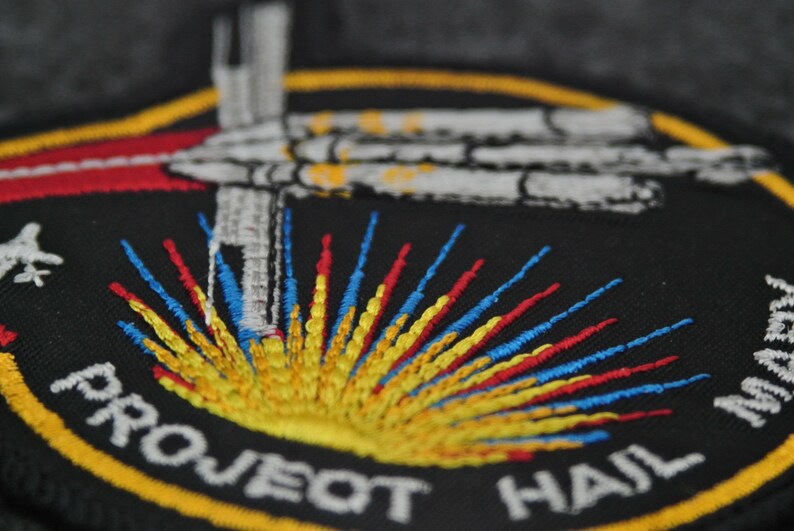 Puede incluir: Parche bordado con fondo negro, con un dise&ntilde;o de nave espacial y el texto "PROJECT HAIL MARS". El parche tiene un borde amarillo y detalles coloridos en rojo, azul y amarillo.