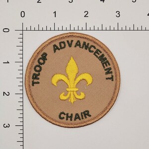 May include: A tan round patch with green embroidered text that reads "Troop Advancement Chair" and a yellow embroidered fleur-de-lis in the center.