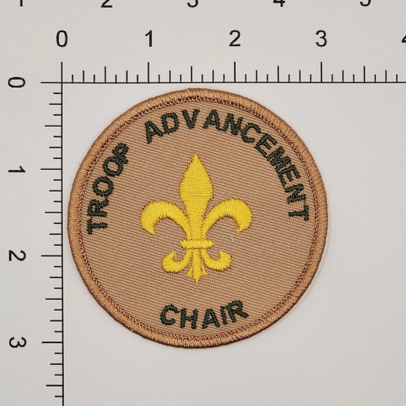 May include: A tan round patch with green embroidered text that reads "Troop Advancement Chair" and a yellow embroidered fleur-de-lis in the center.