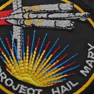 Puede incluir: Parche bordado con fondo negro, con una nave espacial estilizada y una explosi&oacute;n de rayos amarillos, rojos y azules. El texto "PROJECT HAIL MARY" est&aacute; arqueado alrededor del dise&ntilde;o en letras blancas, con un borde amarillo.