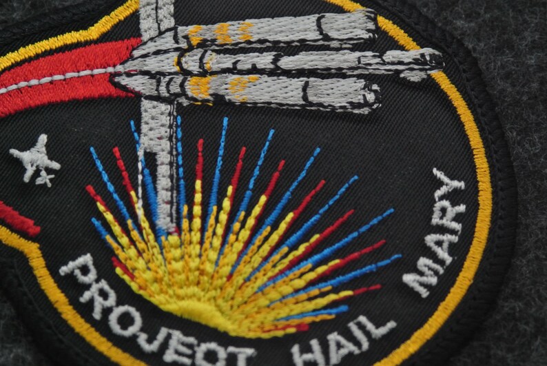 Puede incluir: Parche bordado con fondo negro, con una nave espacial estilizada y una explosi&oacute;n de rayos amarillos, rojos y azules. El texto "PROJECT HAIL MARY" est&aacute; arqueado alrededor del dise&ntilde;o en letras blancas, con un borde amarillo.