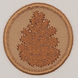 May include: A brown embroidered patch with a brown pine cone design on a beige background.