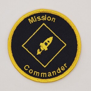 May include: A black and gold embroidered patch with a rocket ship inside a diamond shape. The text "Mission Commander" is embroidered around the diamond.