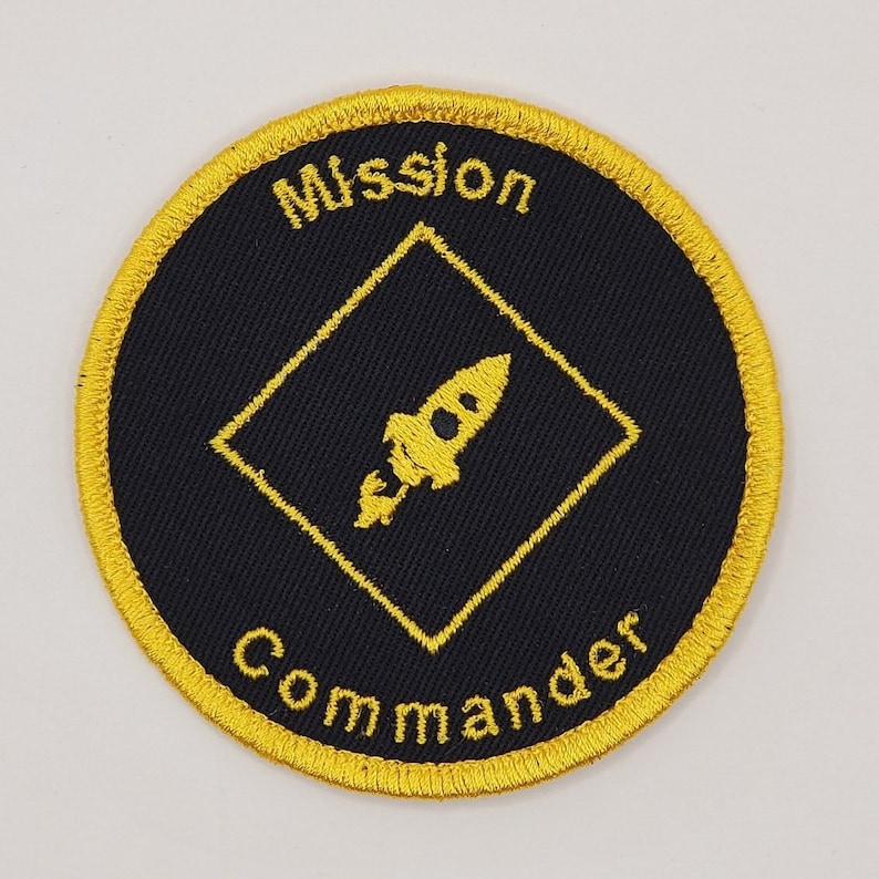 May include: A black and gold embroidered patch with a rocket ship inside a diamond shape. The text "Mission Commander" is embroidered around the diamond.
