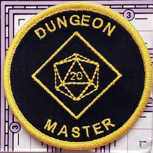 May include: A black and gold embroidered patch with a 20-sided die in the center and the text "DUNGEON MASTER" around the edge.