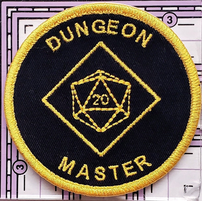 May include: A black and gold embroidered patch with a 20-sided die in the center and the text "DUNGEON MASTER" around the edge.