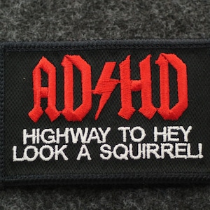 May include: Black rectangular patch with red embroidered text "ADHD" and a lightning bolt, above "HIGHWAY TO HEY LOOK A SQUIRREL!" in white. The patch has a black border and is set against a gray background.